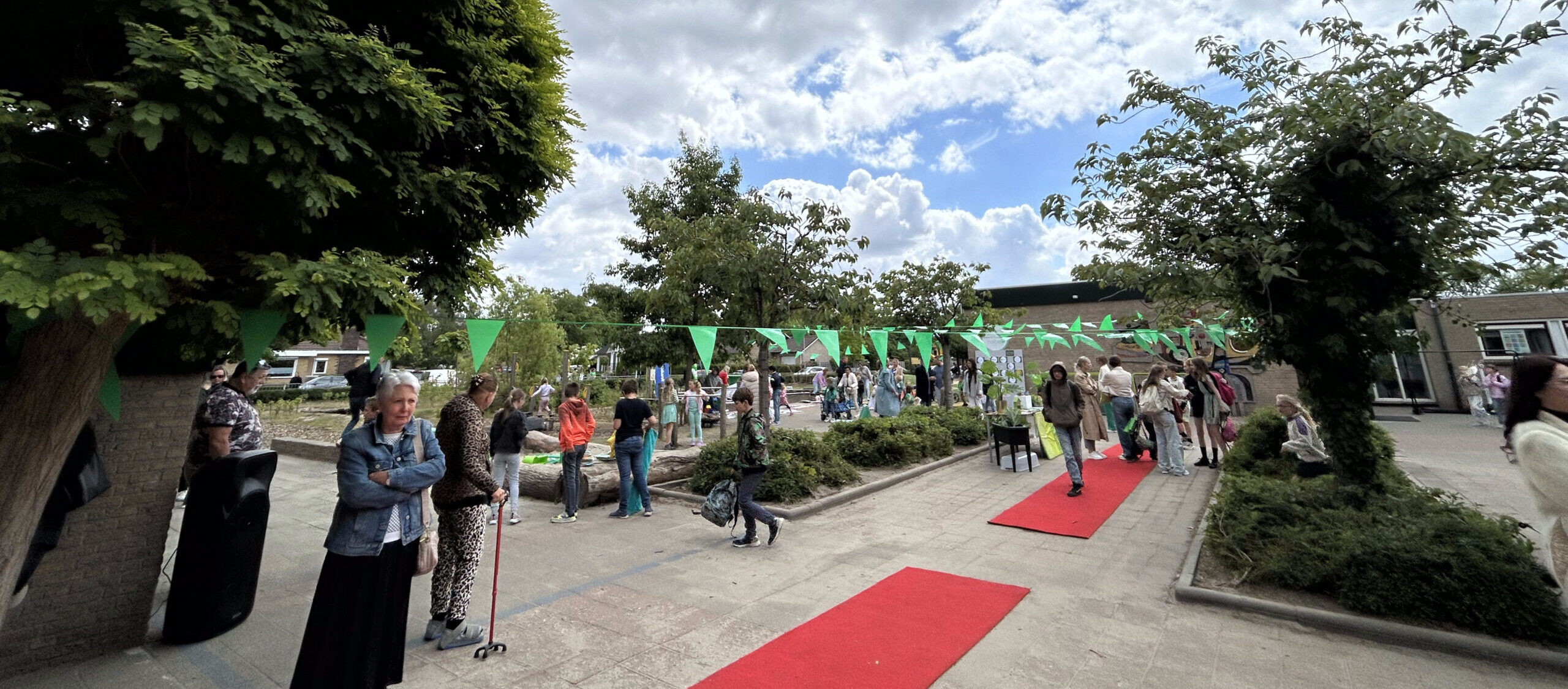 Buitenfeest met groene vlaggetjes, rode lopers en mensen die rondlopen bij bomen en struiken.