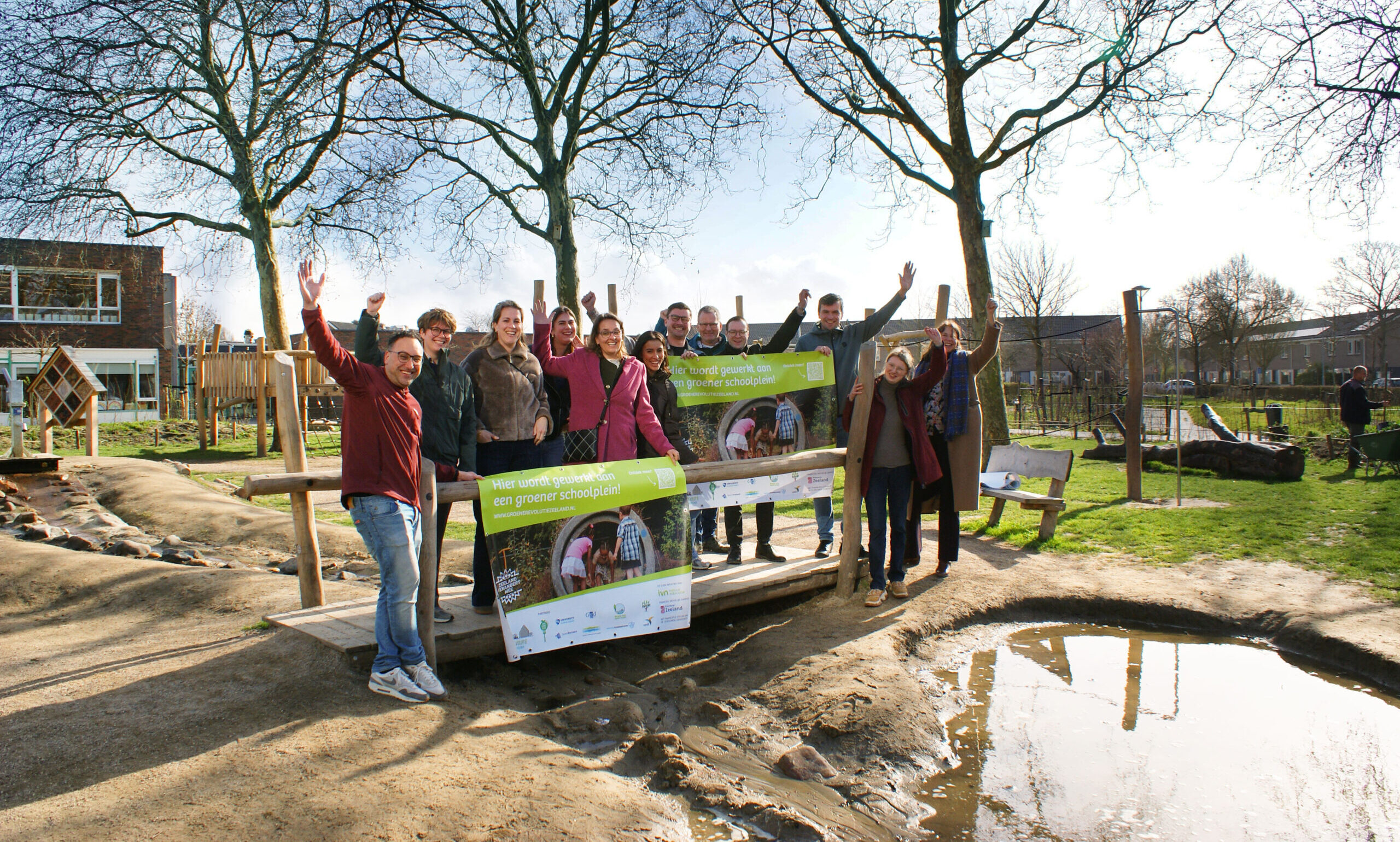 Groep mensen viert de bouw van een groener schoolplein bij een speeltuin met spandoeken en bomen.