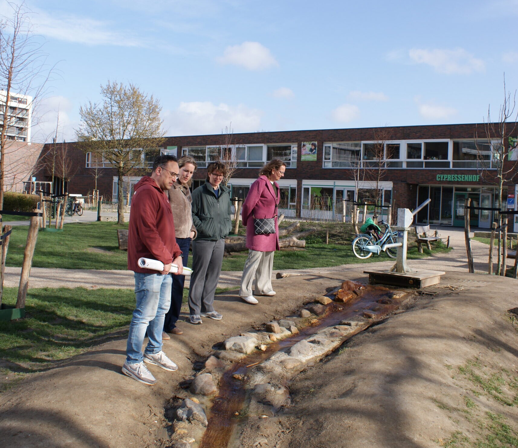 Groep mensen observeert een waterstroom naast een gebouw met fietsen op een zonnige dag.