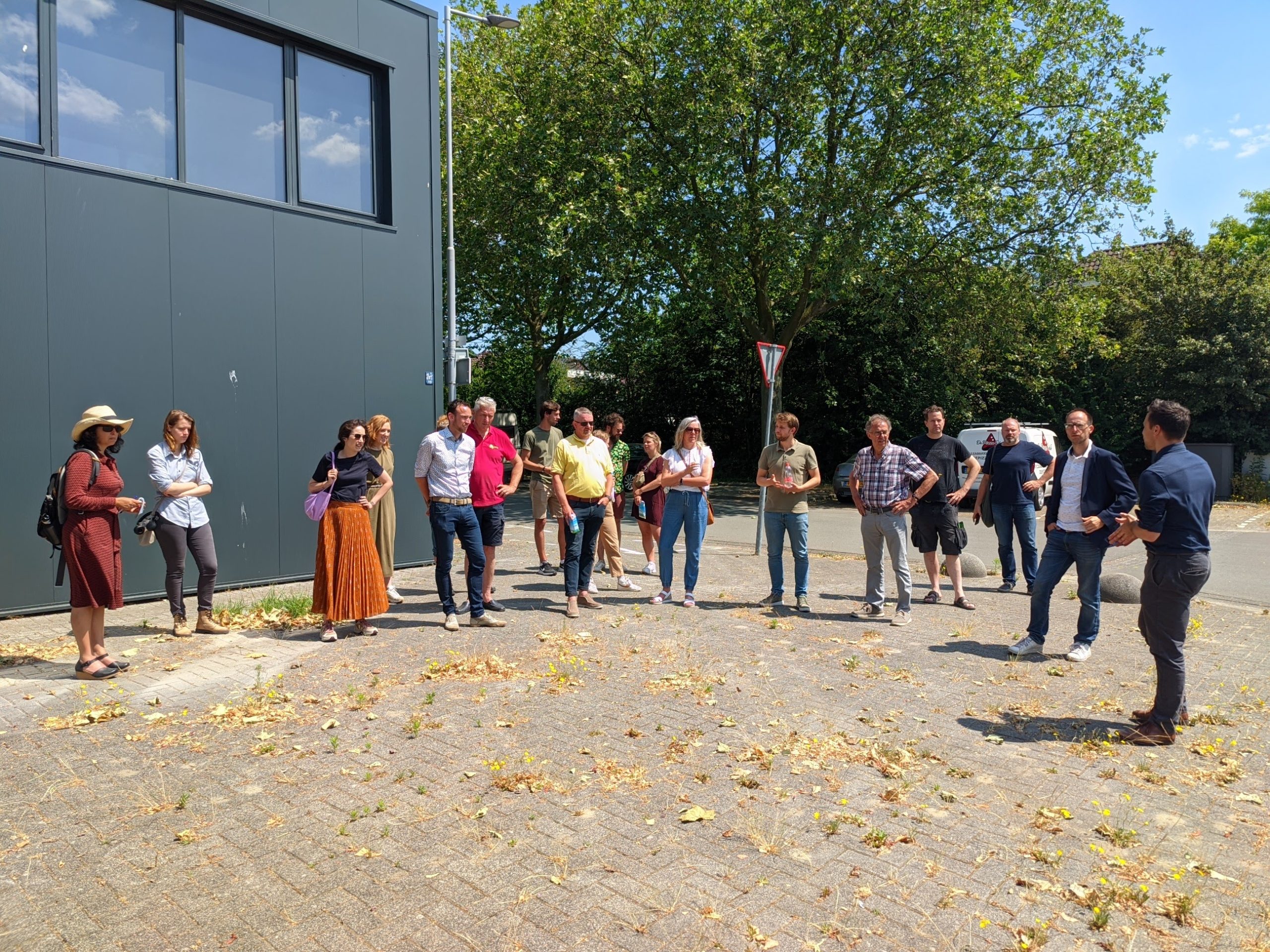 Groep mensen staat buiten bij een gebouw, luistert naar een spreker, zonnige dag.