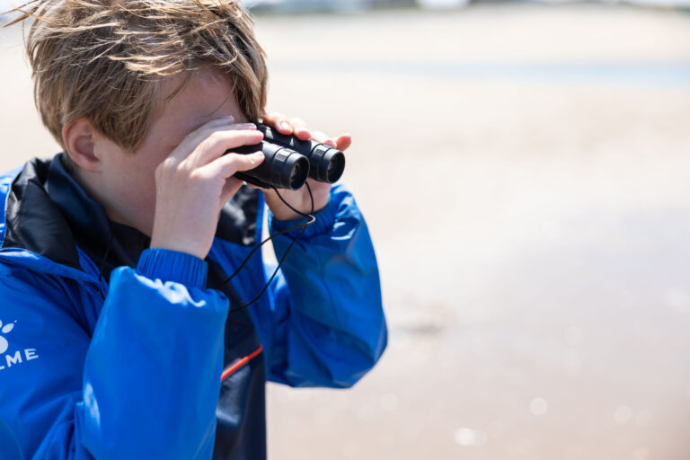 Jongen met blauwe jas kijkt door verrekijker op een zonnig, winderig strand.