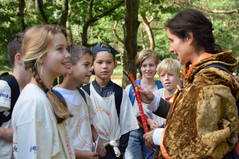 Een vrouw in middeleeuwse outfit spreekt met een groep kinderen in een bos.