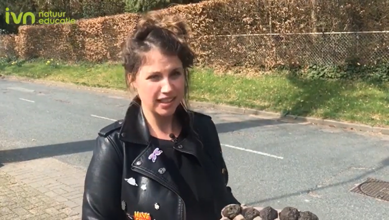 Een vrouw in een leren jas staat buiten met een blad, omgeven door een groene omgeving.