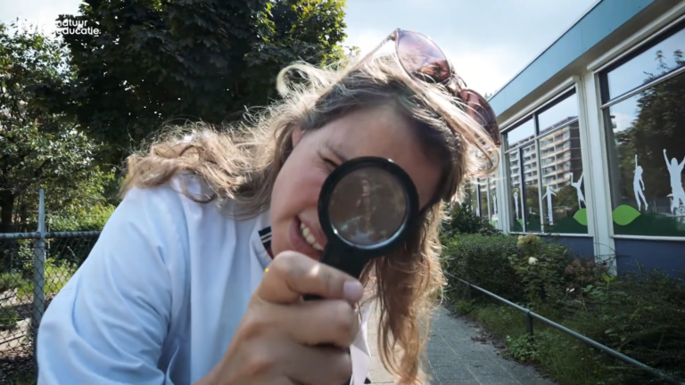 Vrouw met vergrootglas buigt zich voorover bij een schoolgebouw met bomen op de achtergrond.