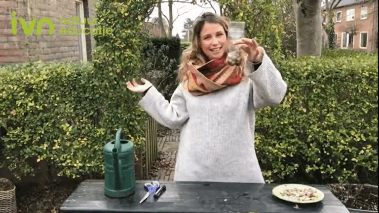 Vrouw in tuin met grijze trui houdt fles omhoog. Op tafel een gieter, schaar en zaden.