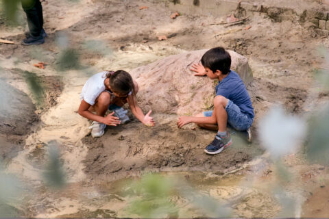 Kinderen spelen in modder bij een grote rots, omgeven door natuurlijk terrein.