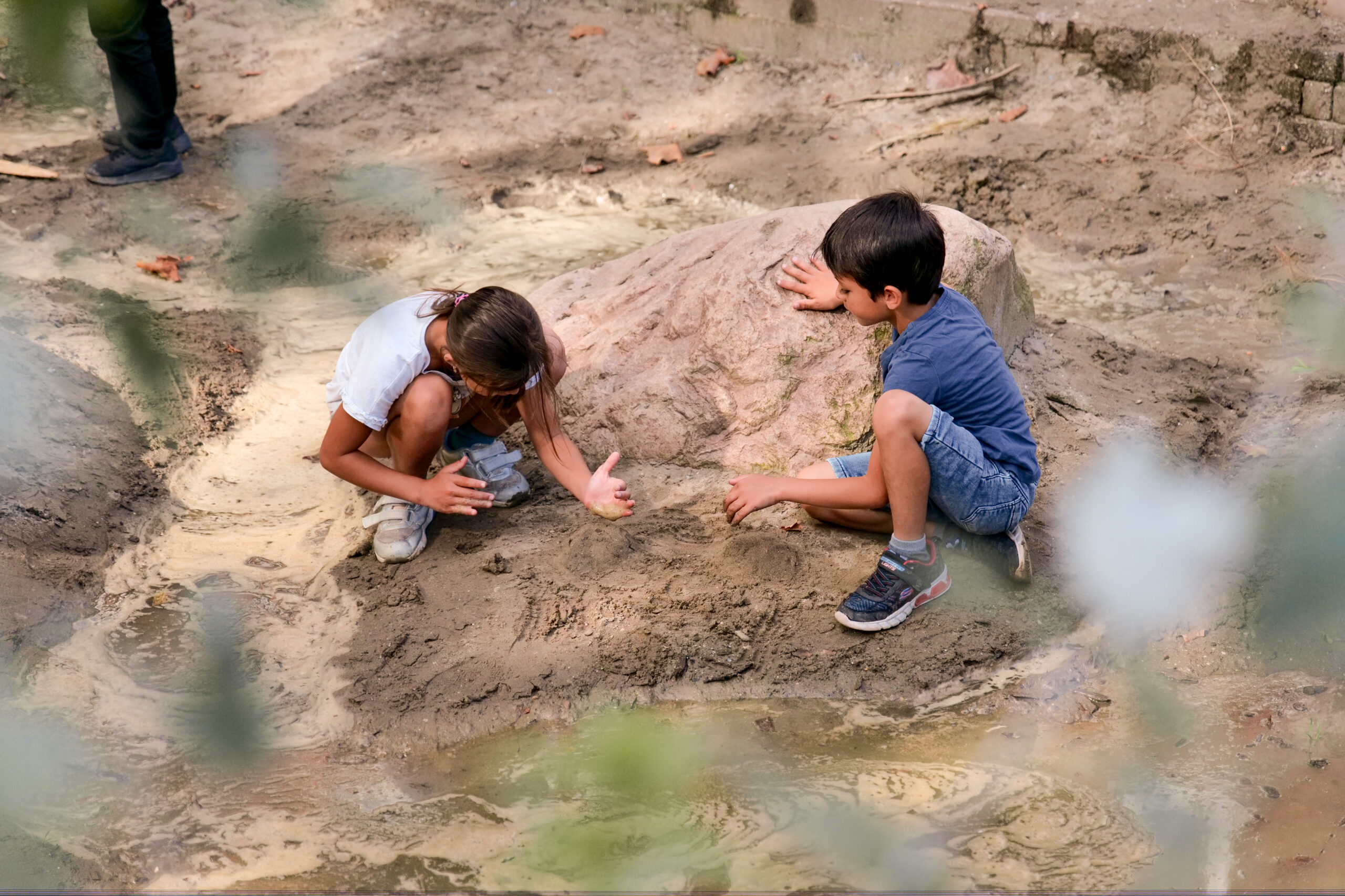 Kinderen spelen in modder bij een grote rots, omgeven door natuurlijk terrein.