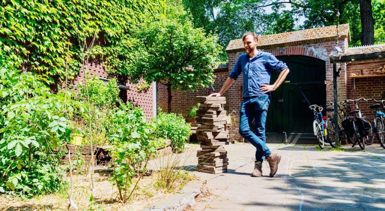 Man leunt op stapel stenen in een groene tuin met fietsen en een stenen muur.