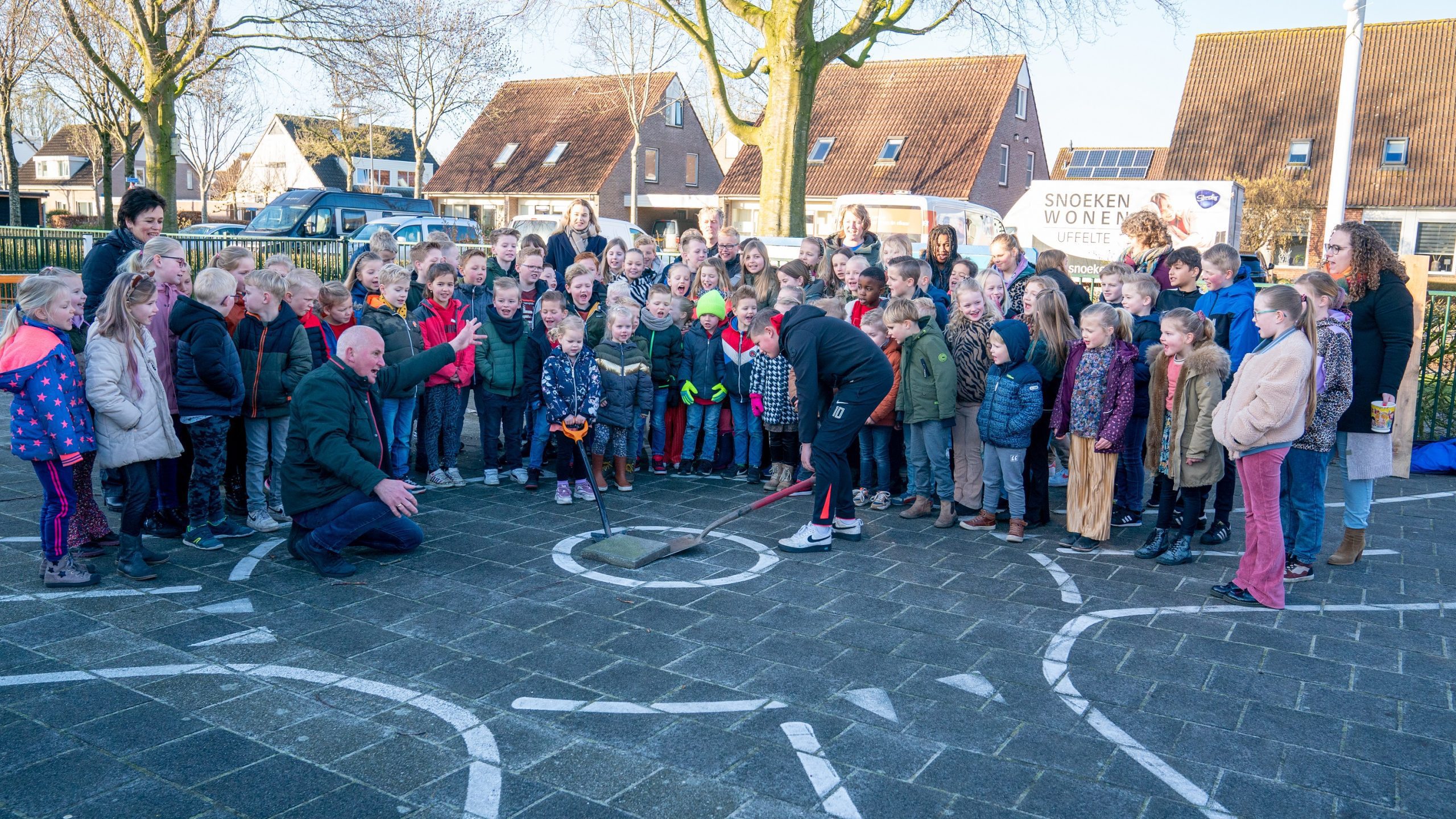Een groep kinderen en volwassenen kijkt naar een man terwijl hij iets op de grond opent.