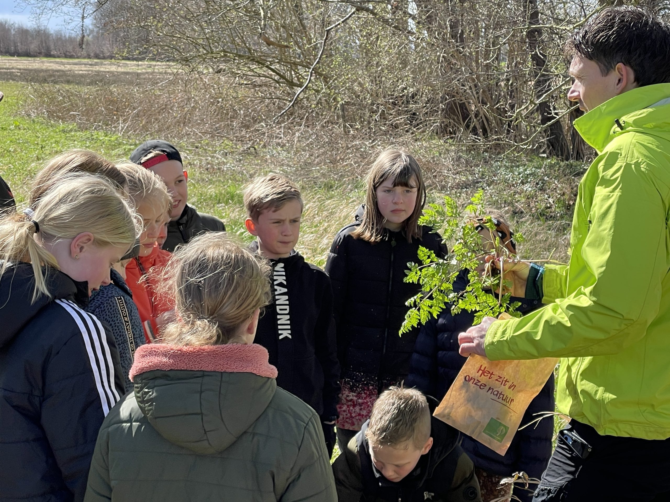 Een groep kinderen observeert planten die door een man in gele jas worden getoond.