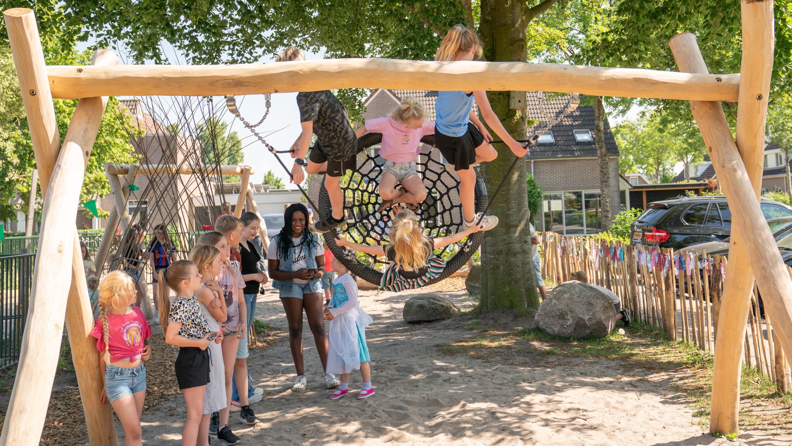 Kinderen spelen op een nestschommel in een zonnige speeltuin met bomen en een houten hek.