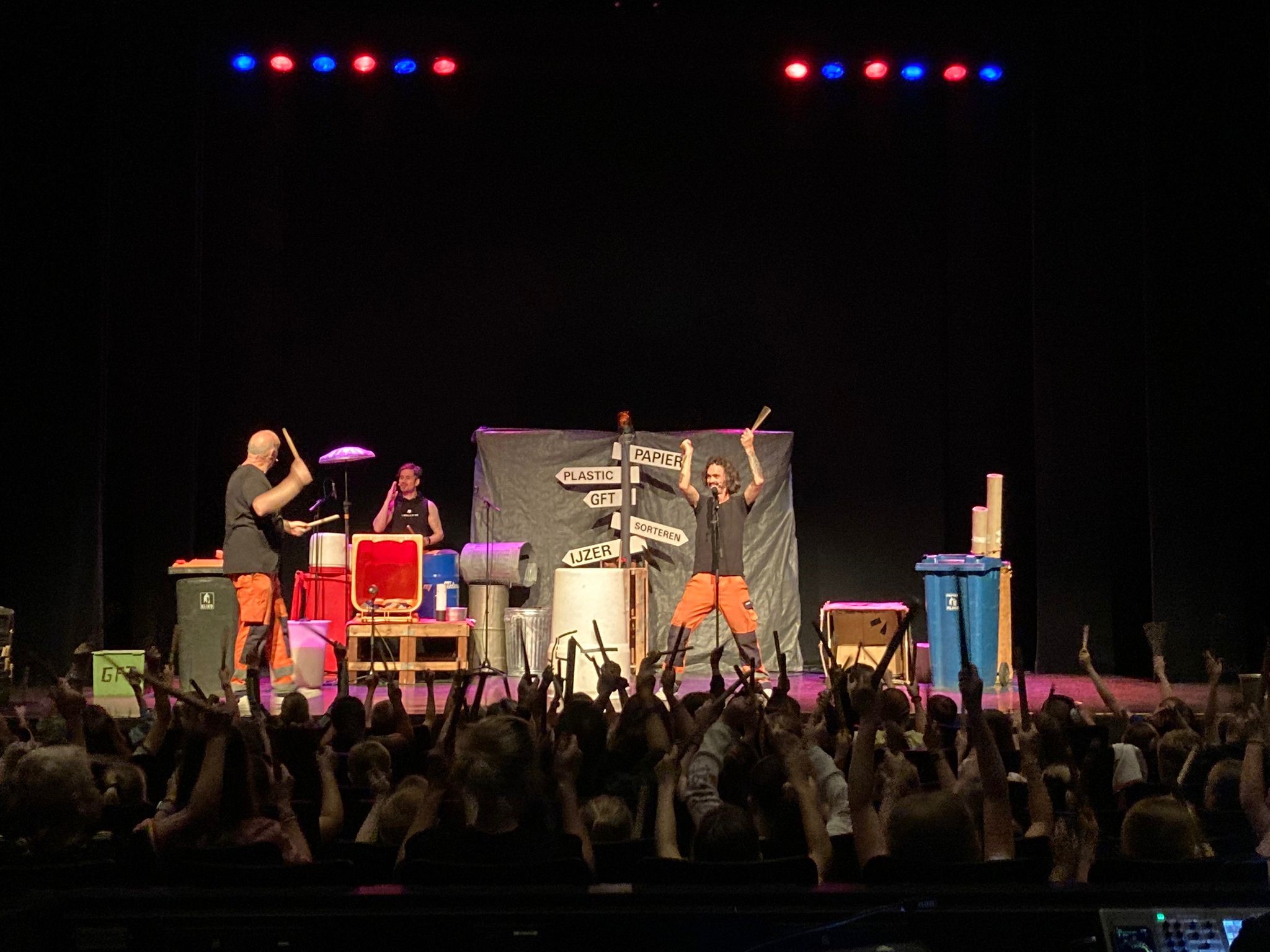 Twee muzikanten drummen op gerecycleerde voorwerpen op een podium voor een enthousiast publiek.