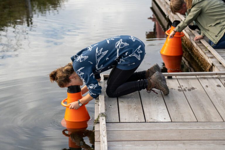Twee personen kijken in het water met gebruik van oranje kijkers vanaf een houten steiger.
