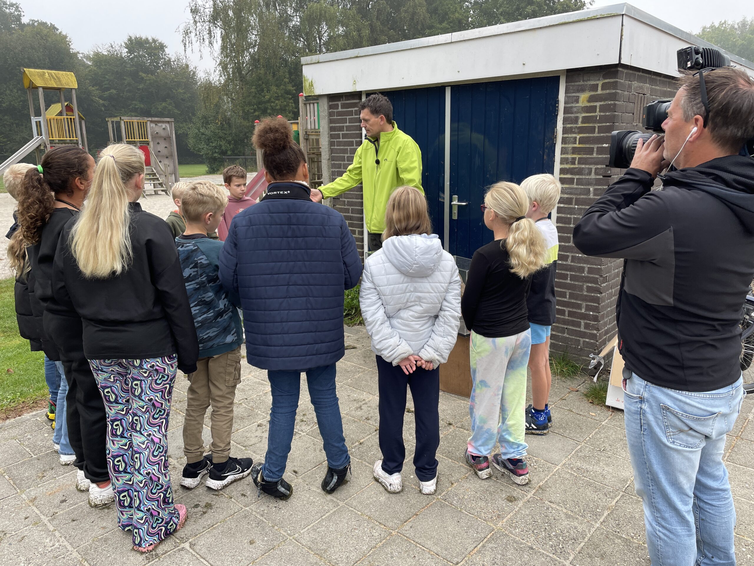 Een groep kinderen luistert naar een man voor een blauw gebouw, terwijl een cameraman filmt.