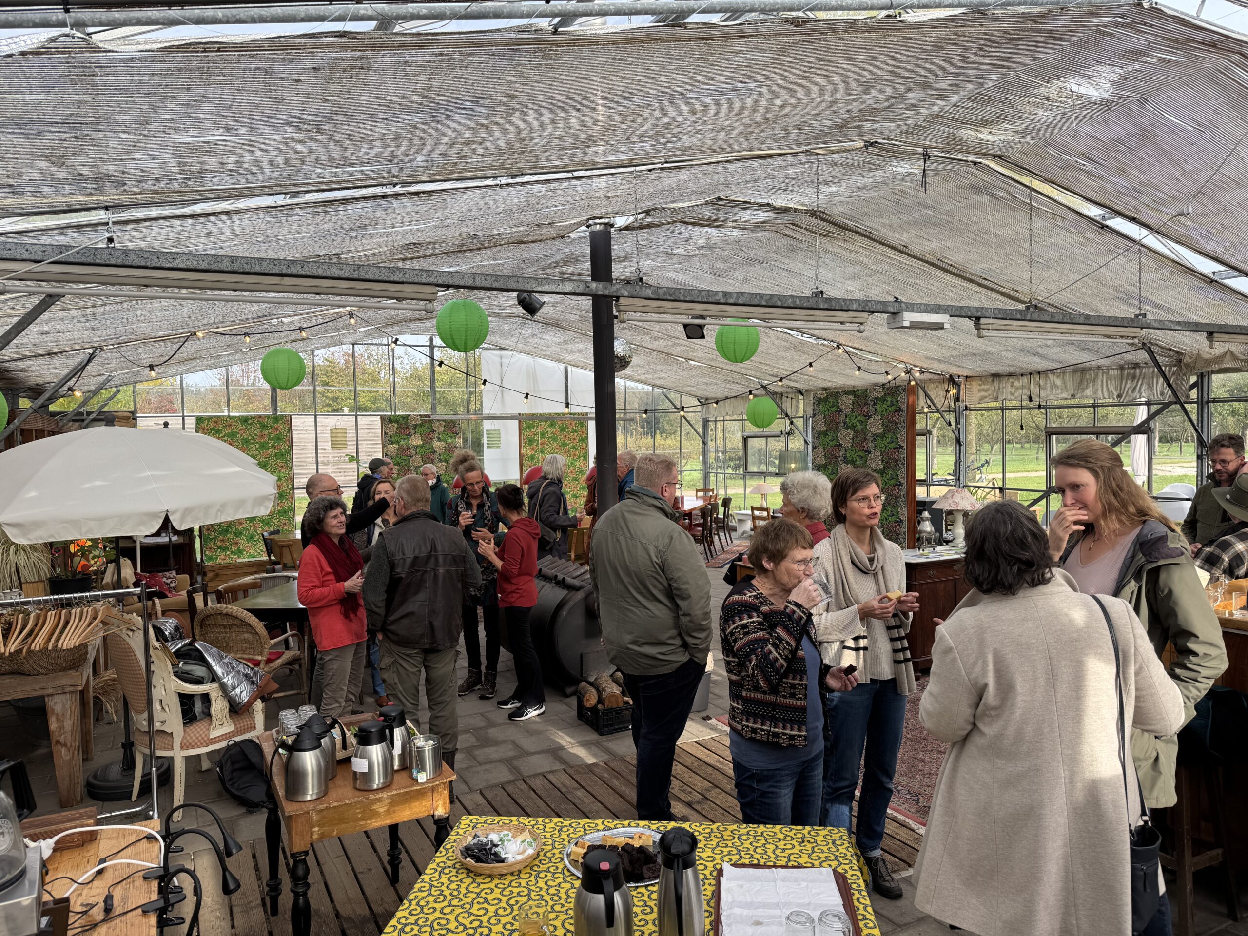 Groep mensen in een kas, pratend en genietend van drankjes, met groene lantaarns aan het plafond.