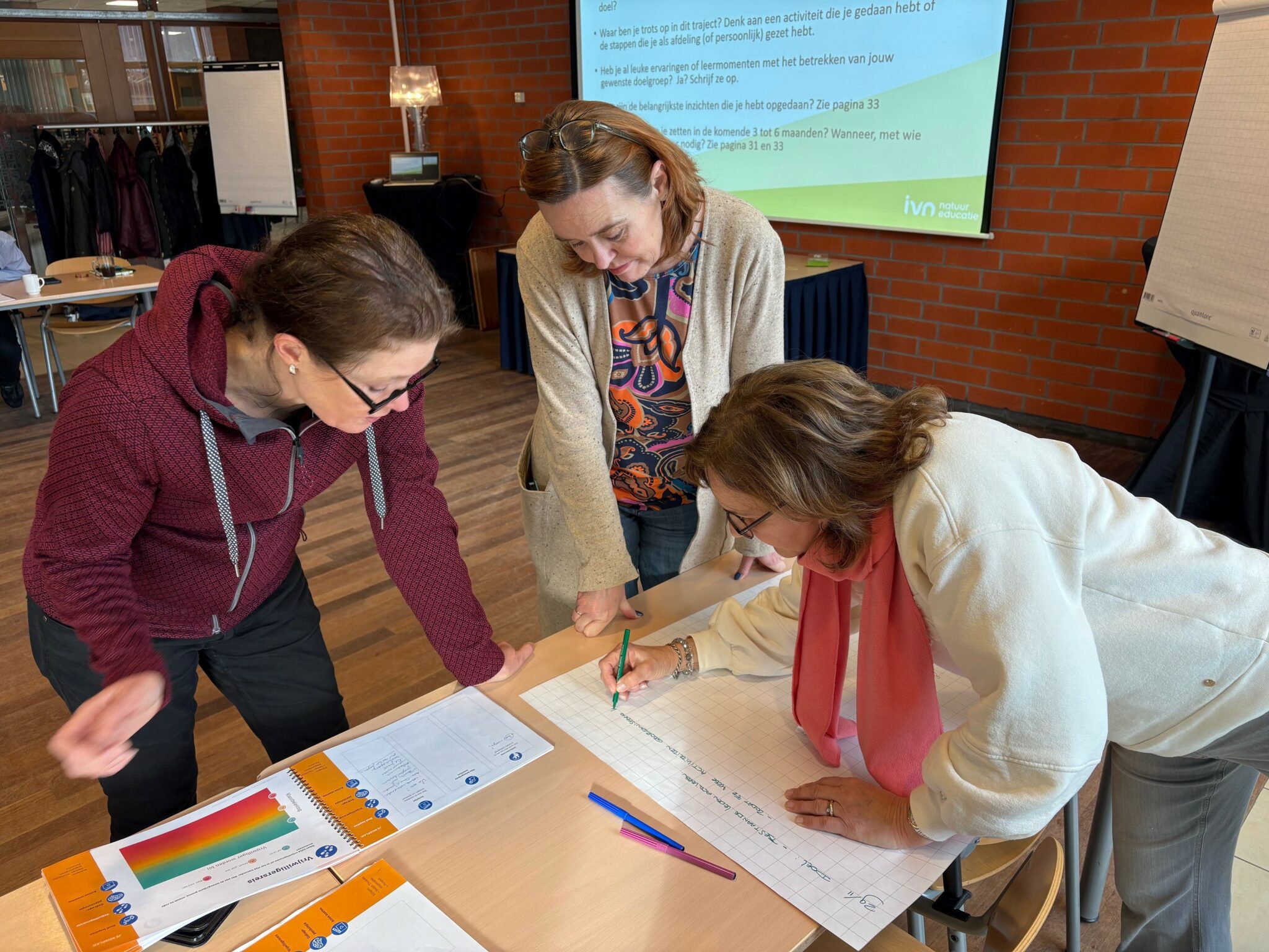 Drie mensen werken samen aan een groot vel papier tijdens een bijeenkomst in een vergaderruimte.