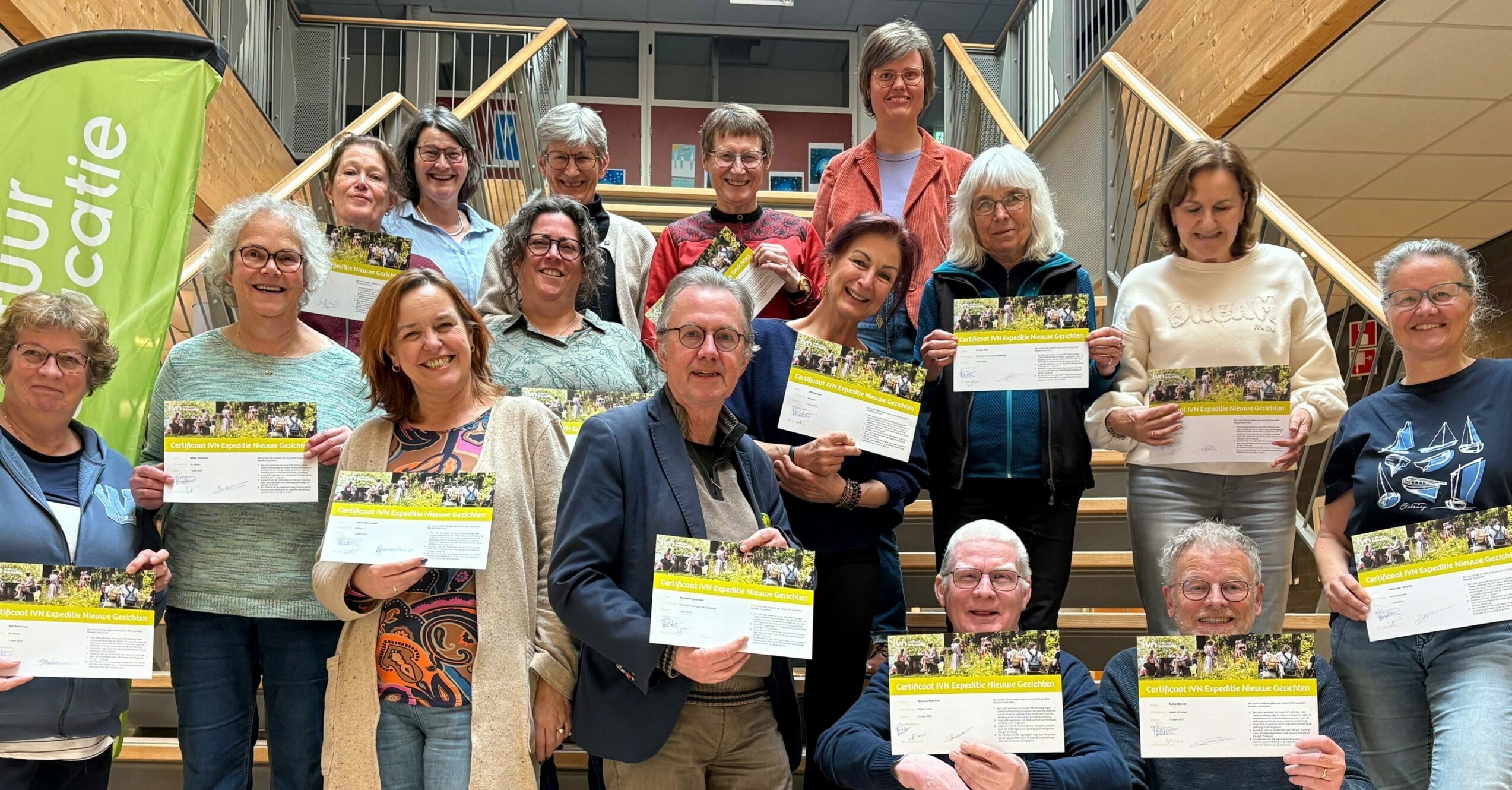 Groep mensen poseert met certificaten op een trap, naast een groene vlag.