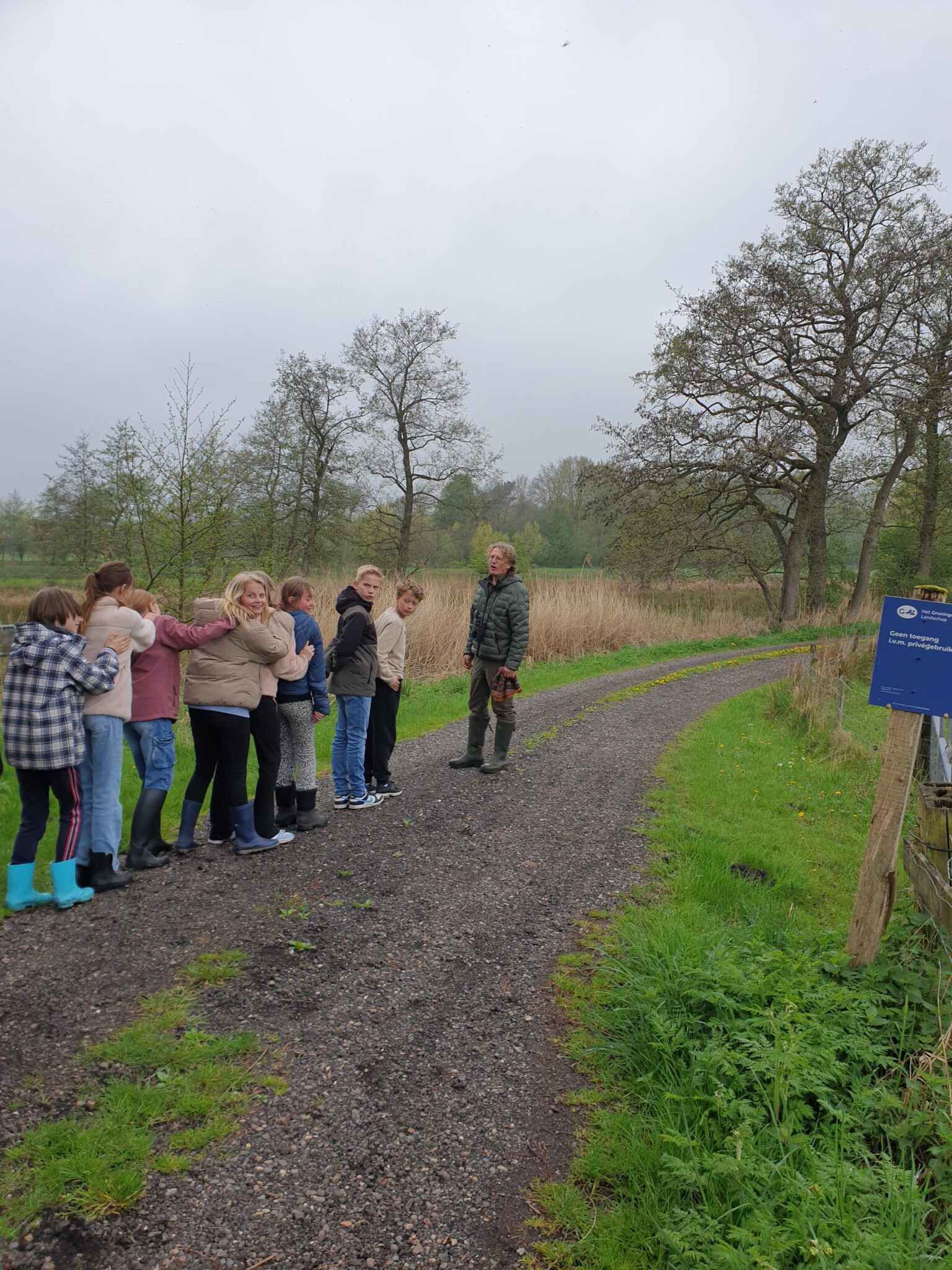 Groep kinderen en volwassene op landweg, omgeven door bomen en gras, naast informatiebord.