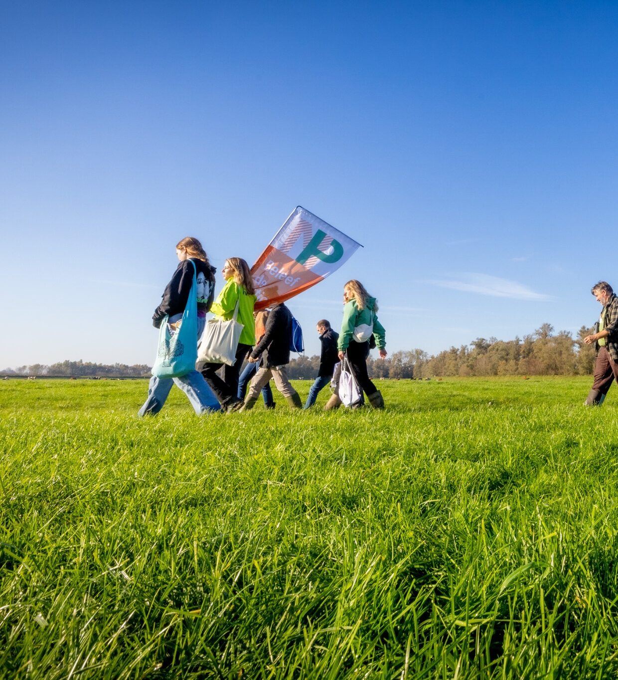 Groep mensen loopt over een grasveld onder een heldere blauwe lucht, met een vlag.