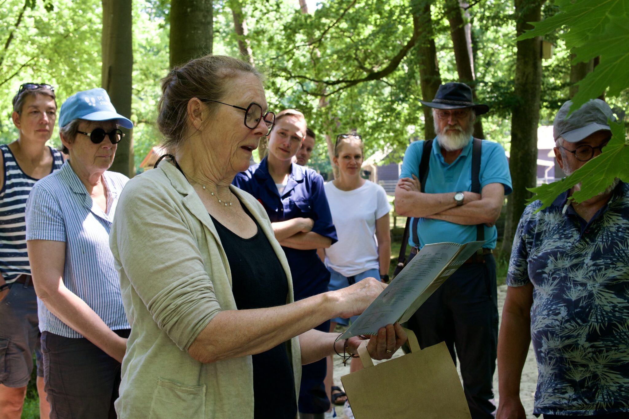 Groep mensen luistert naar vrouw die buiten voorleest, omgeven door bomen.