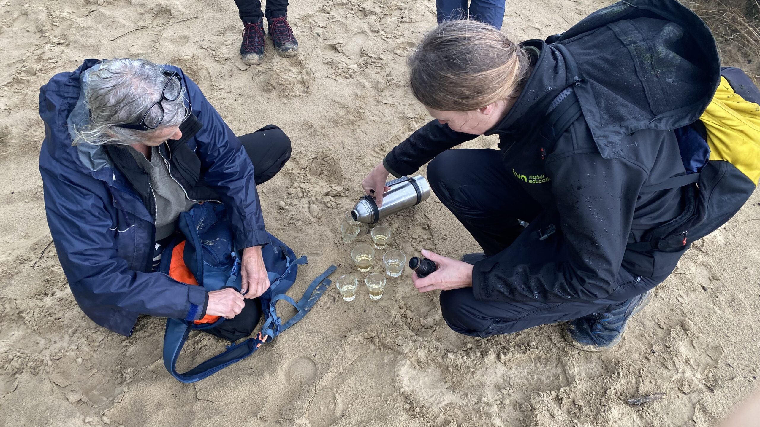 Twee mensen op zand, één schenkt drankjes uit een thermosfles, glazen staan op de grond.