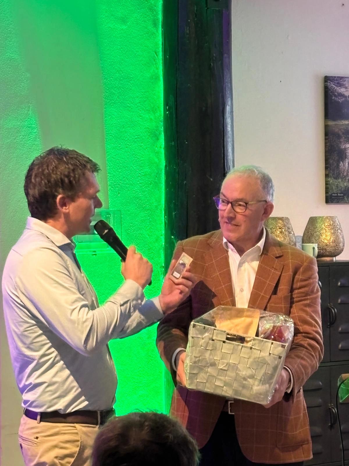 Twee mannen bij een evenement; een houdt een microfoon, de ander een cadeaupakket vast.