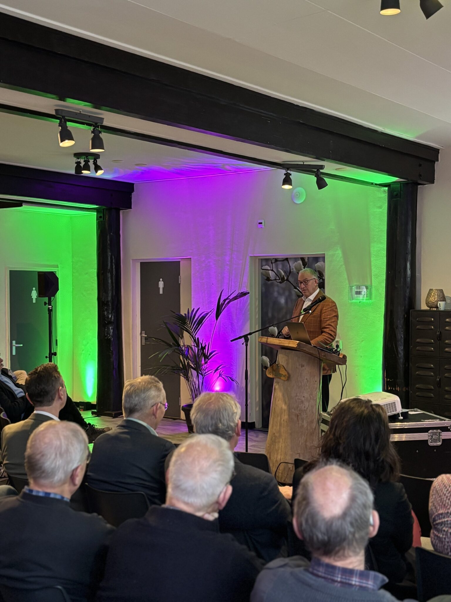 Een persoon spreekt voor een klein publiek in een zaal met kleurrijke verlichting.