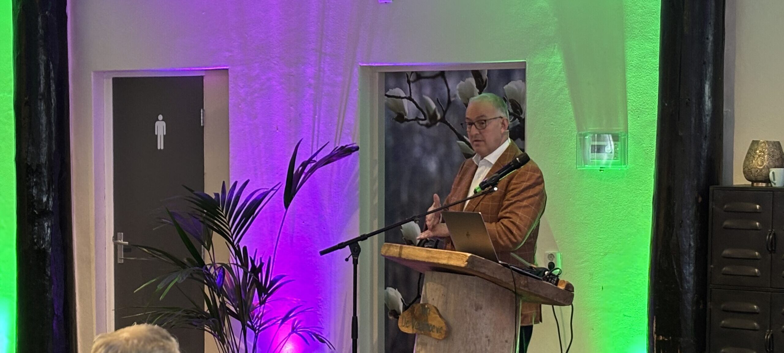 Man geeft presentatie aan publiek in verlichte ruimte met groene planten.