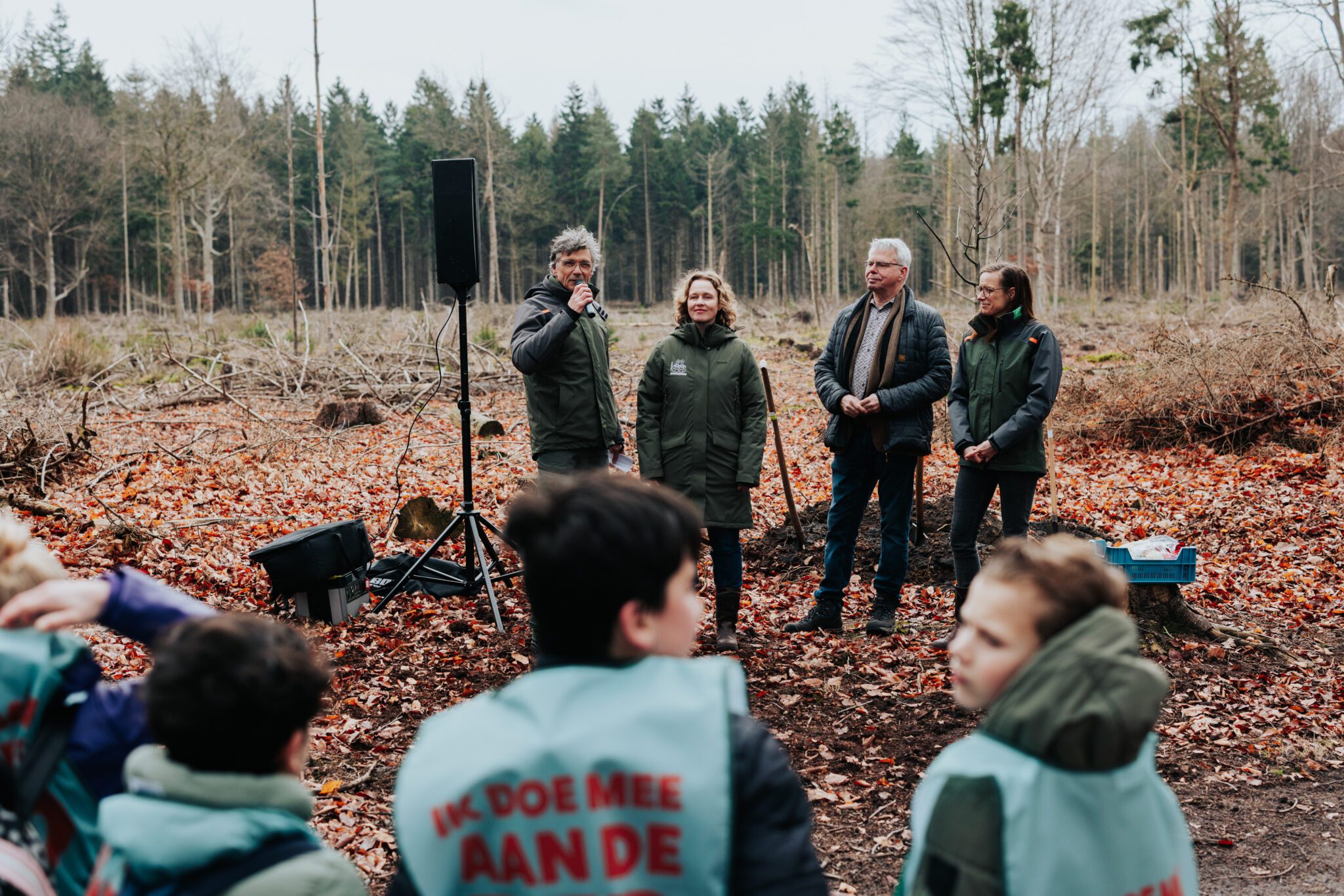 Mensen staan buiten in een herfstbos, luisterend naar een spreker bij een microfoon.