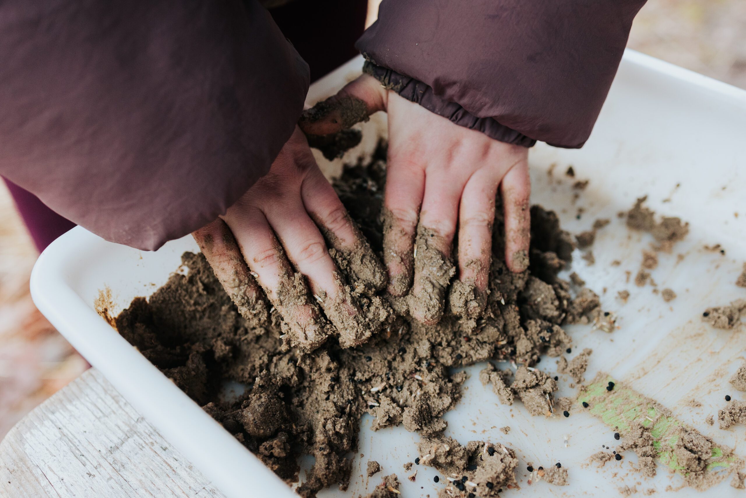 Handen kneden natte aarde in een witte bak, met een donkere jas aan.
