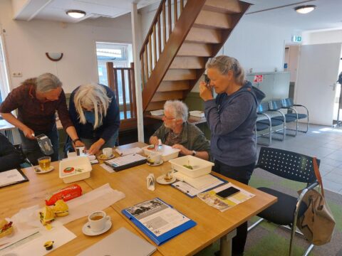 Groep mensen onderzoekt planten aan een tafel met koffiekopjes en papieren in een lichte kamer.