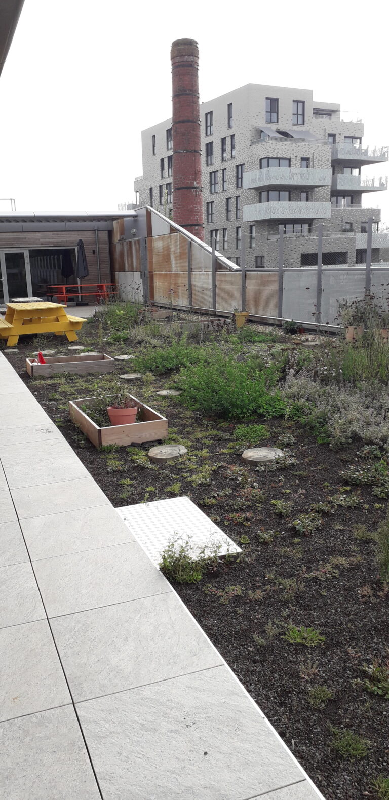 Daktuin met plantenbakken, geel picknickbankje, industrieel gebouw en schoorsteen op de achtergrond.