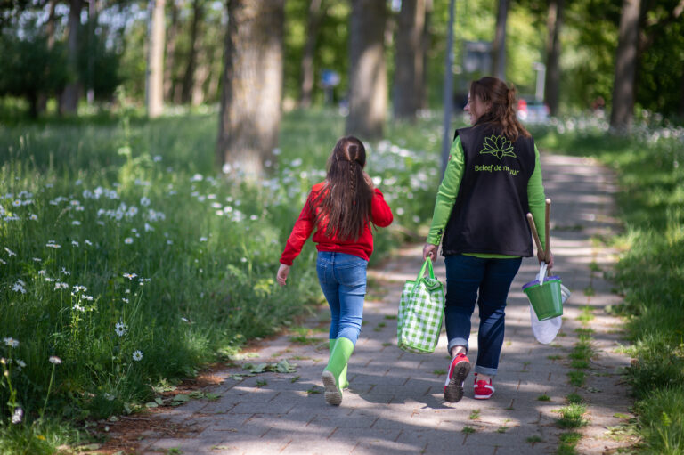 Twee personen lopen over een bospad met bloemen, ze dragen tassen en een net.