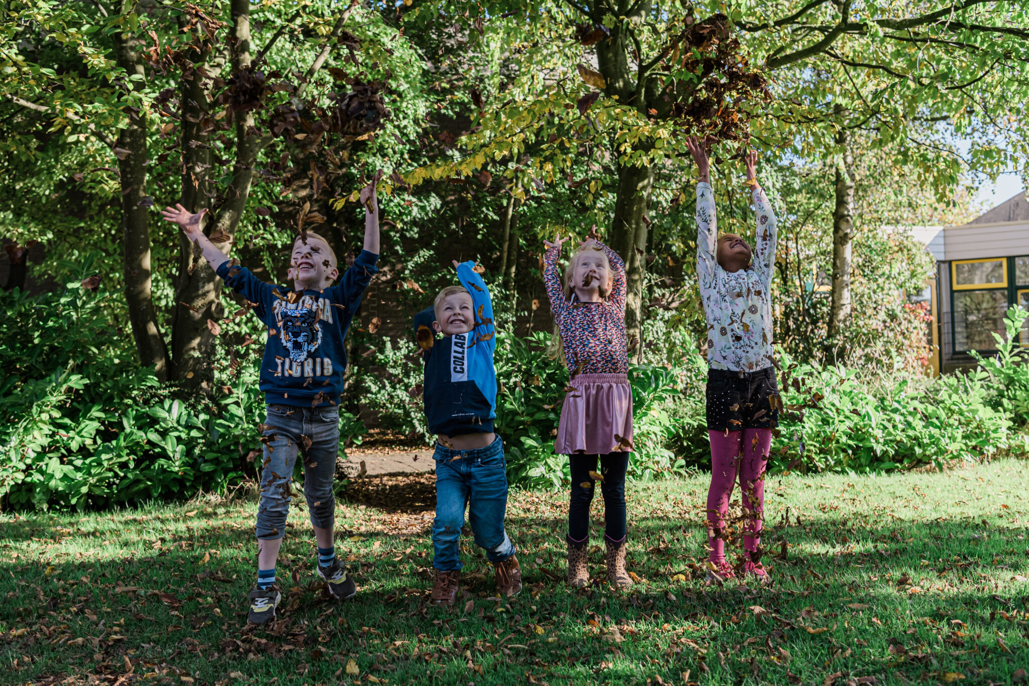 Vier kinderen gooien bladeren omhoog in een groene tuin, stralend van plezier.