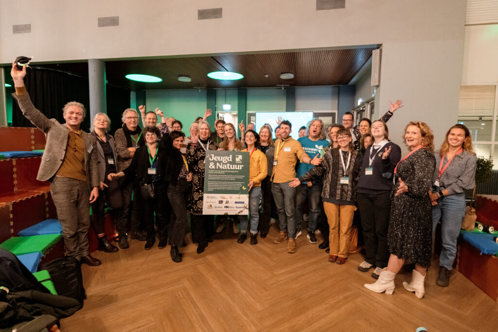 Groep mensen poseert vrolijk met bord "Jeugd & Natuur" in een zaal.