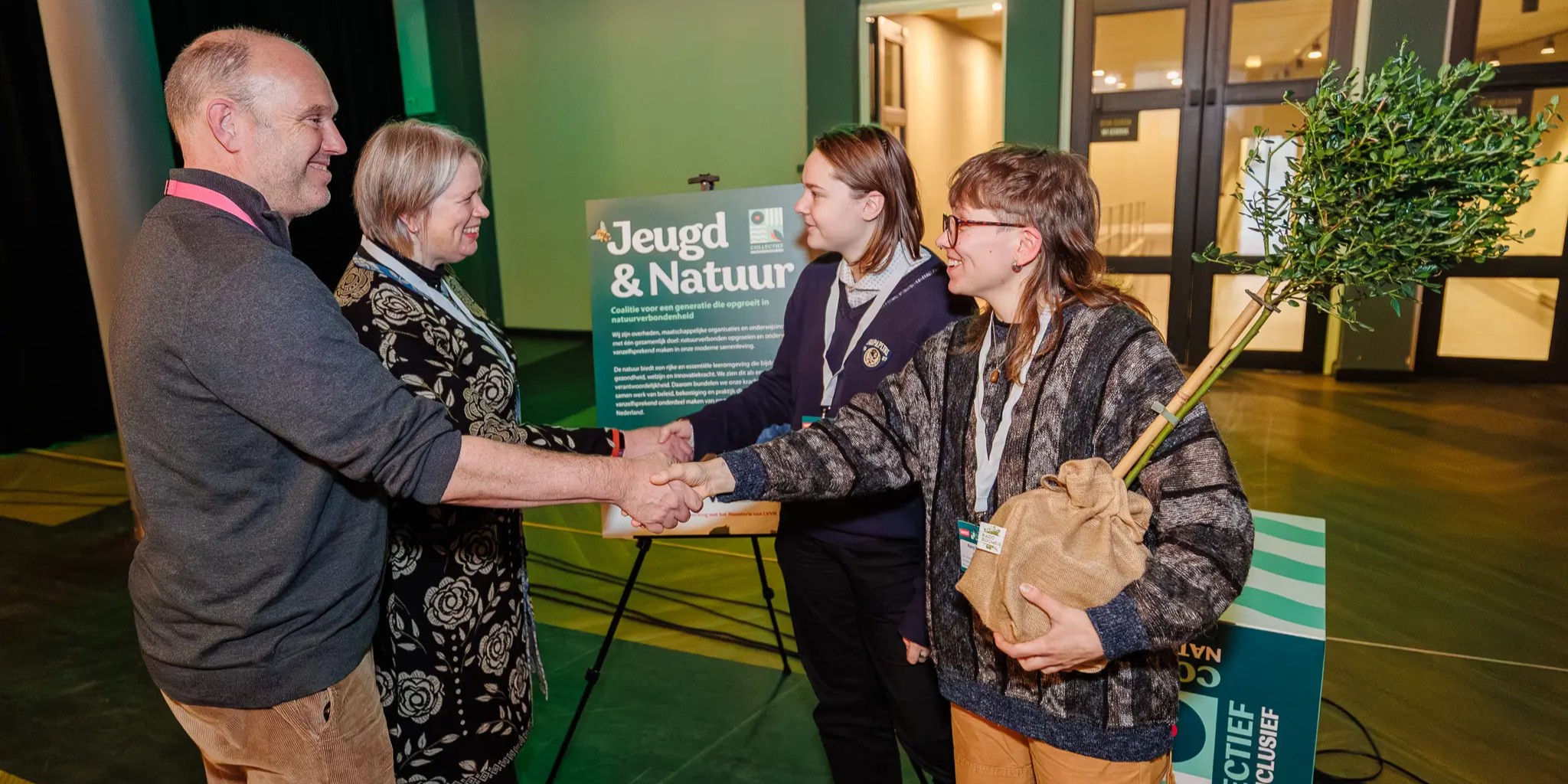 Vier mensen schudden handen, met een bord "Jeugd & Natuur" en een jonge boom in zicht.