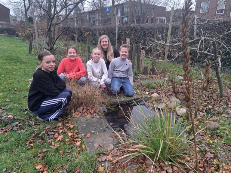 Groep kinderen en een volwassene bij een kleine vijver in een herfstachtig park.