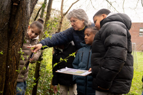 Een vrouw wijst kinderen op planten tijdens een buitenles in een bosrijke omgeving.