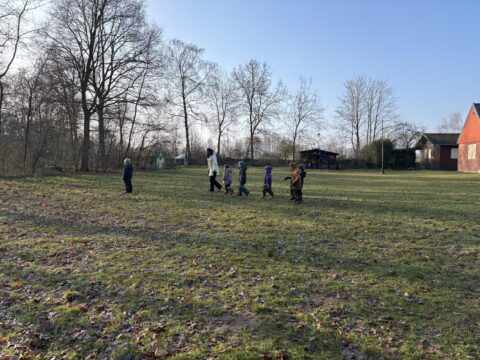 Volwassene en kinderen wandelen over grasveld, omgeven door kale bomen en een gebouw.