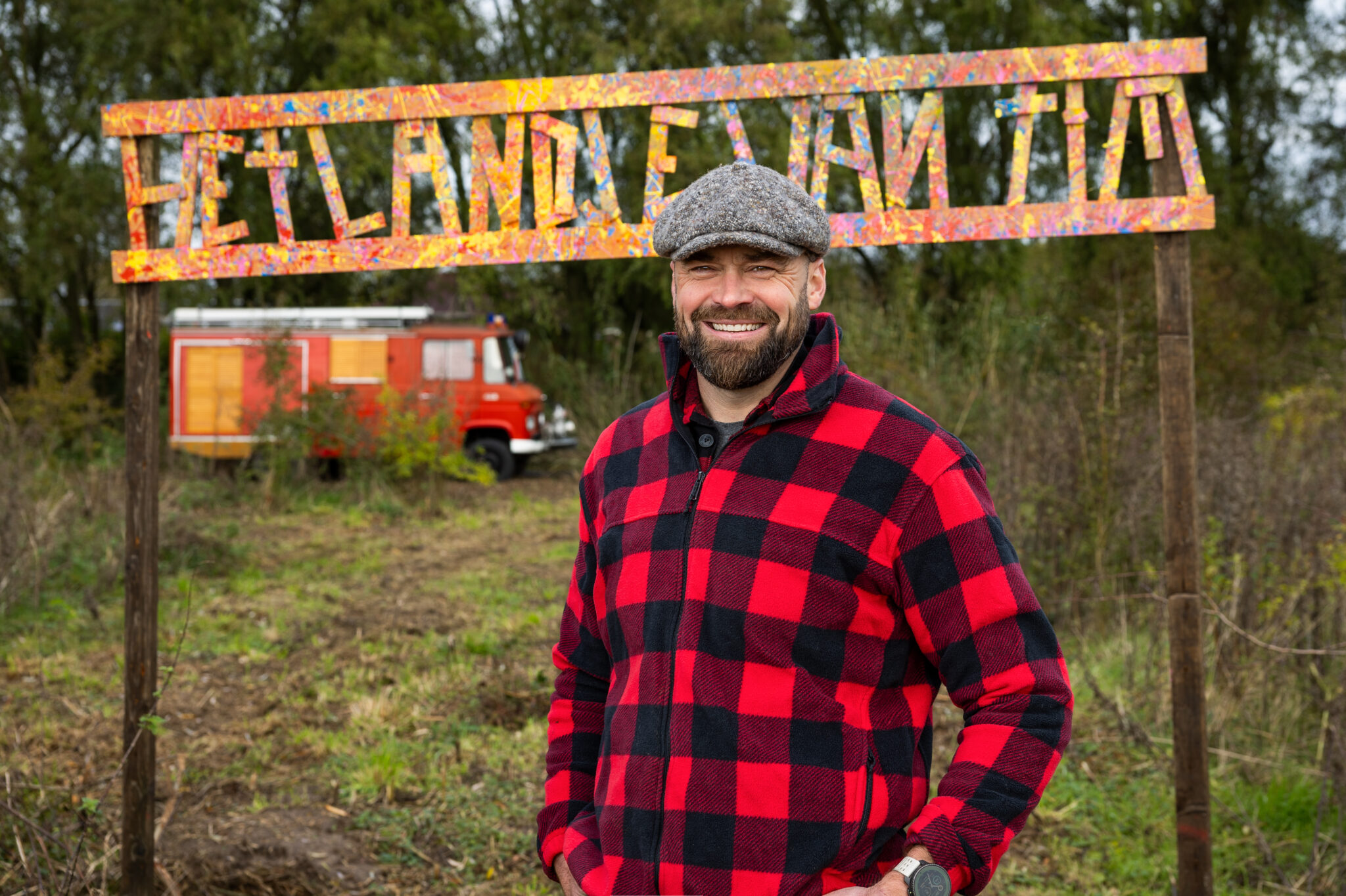 Man met rode houthakkersblouse voor kleurrijk bord "HET LAND VAN AIA" en een rode brandweerauto.
