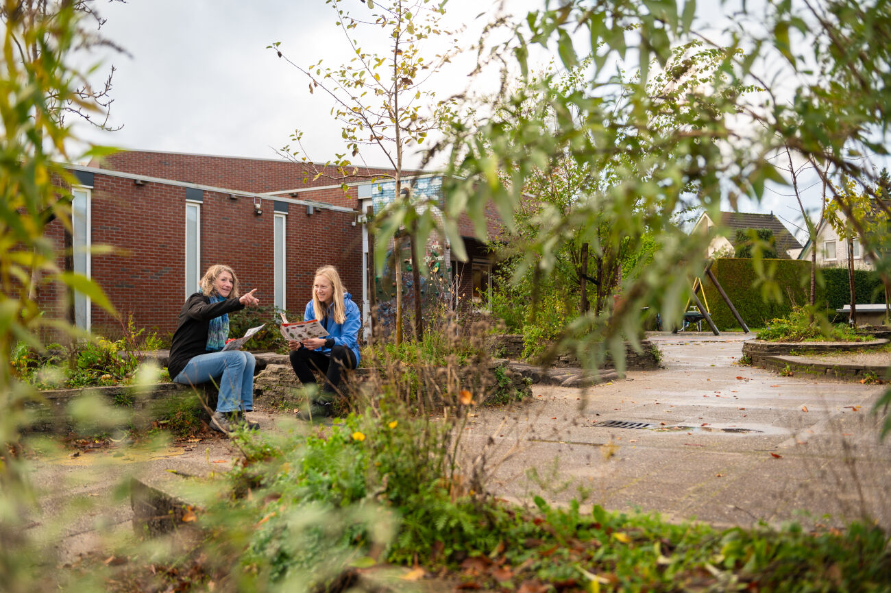 Twee personen praten buiten op een bankje bij een bakstenen gebouw omgeven door planten.
