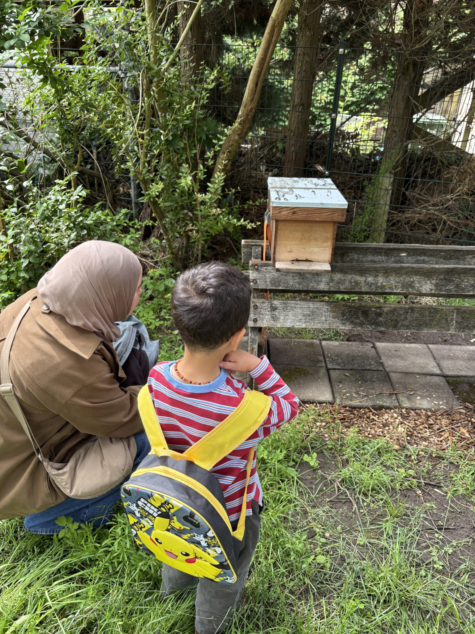 Een vrouw en een kind met een Pikachu-rugzak kijken naar een bijenhuisje in een tuin.