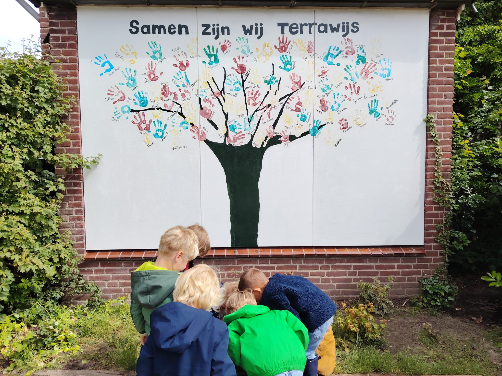 Muur met handafdruk-boom en kinderen ervoor, tekst "Samen Zijn Wij Terrawijs" erboven.