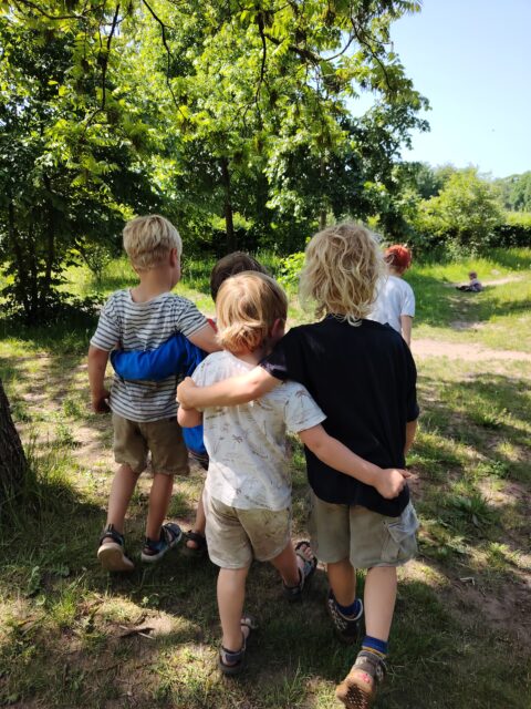 Vier kinderen lopen samen door een zonnig bospad, onder groene bomen.
