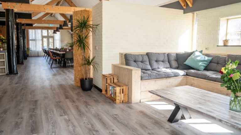Lichte woonkamer met houten vloer, hoekbank, eethoek en planten; veel natuurlijk licht.
