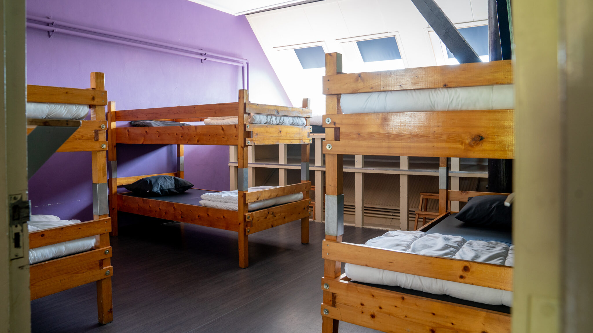 Slaapzaal met houten stapelbedden, paarse muur en dakramen.