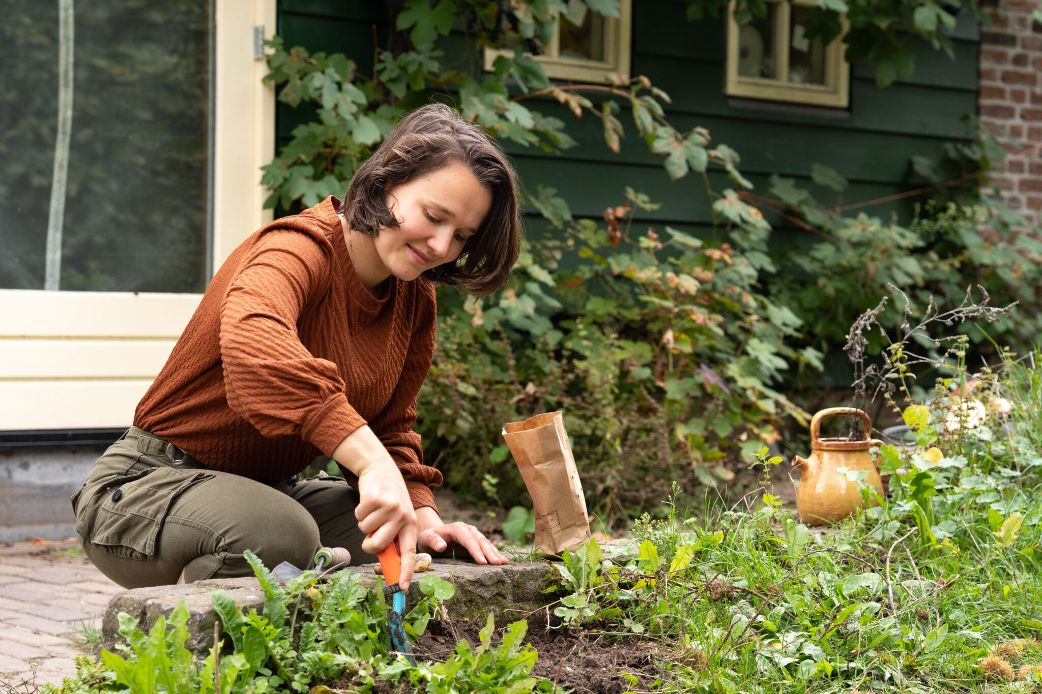 Vrouw in oranje trui tuiniert naast een huis, omgeven door groen en een koperen gieter.