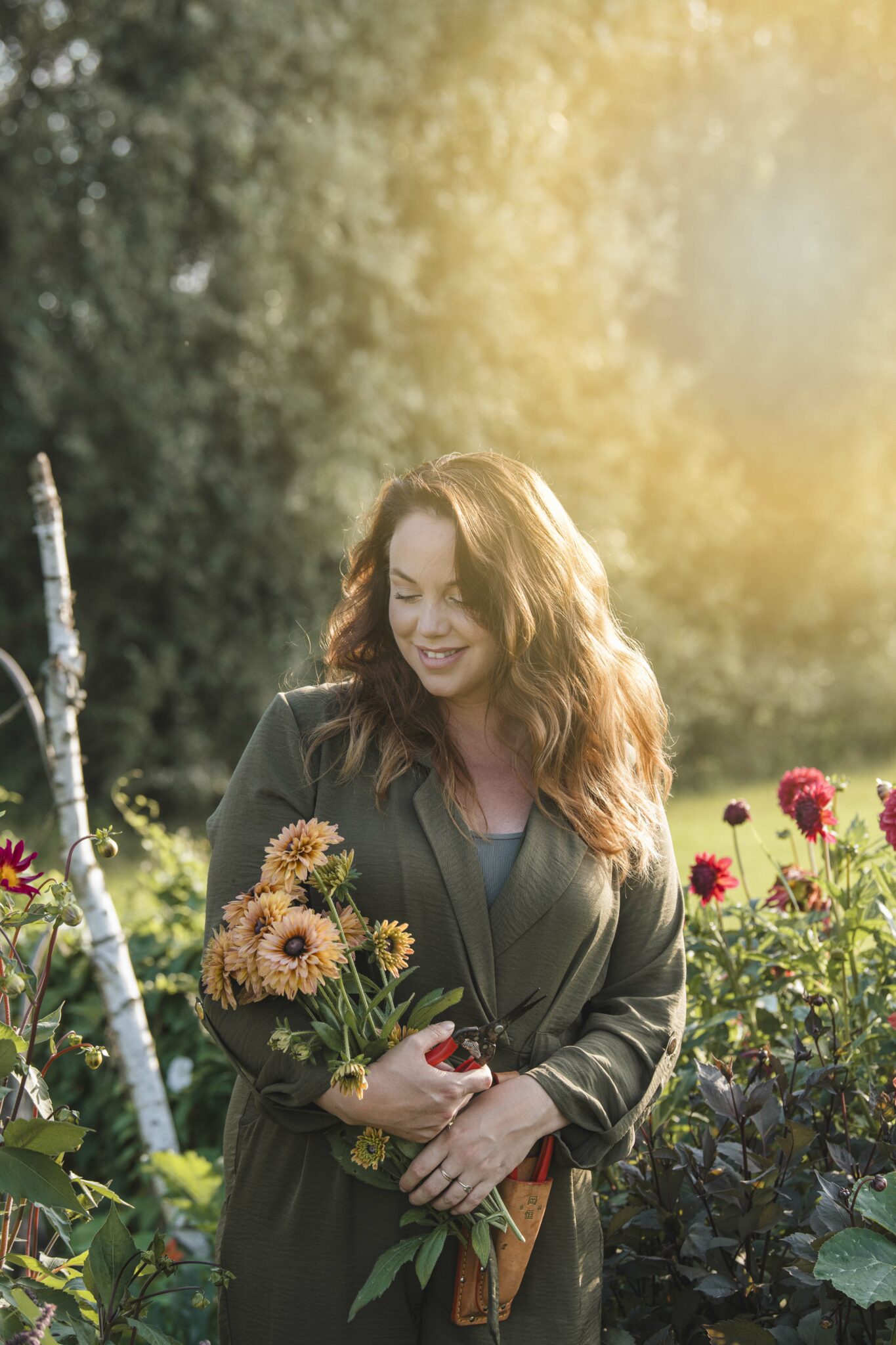 Vrouw in tuin, groene jas, houdt bloemen vast, omringd door bloemen, zonlicht op de achtergrond.