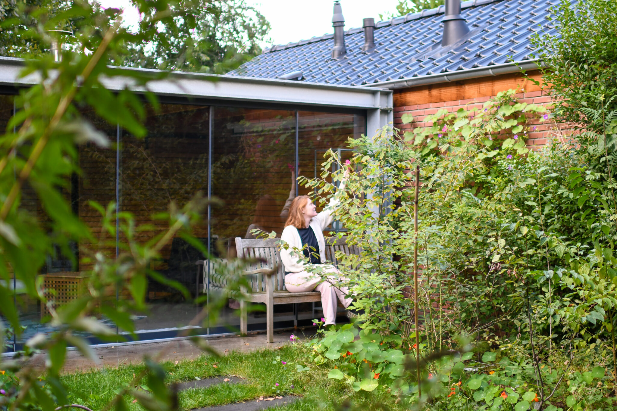 Vrouw zit op bank naast een huis met glazen gevel, omgeven door weelderige tuin.