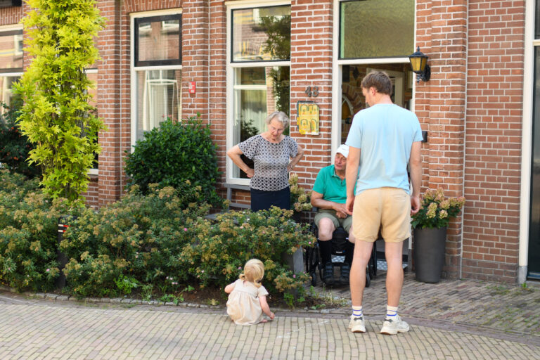 Man staat voor een huis, pratend met een vrouw en man in rolstoel. Kindje speelt op de stoep.